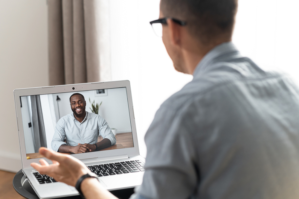 Two mens talking via video connect, an African-American smiles on the laptop screen, chatting online, talk in the distance. View above shoulder