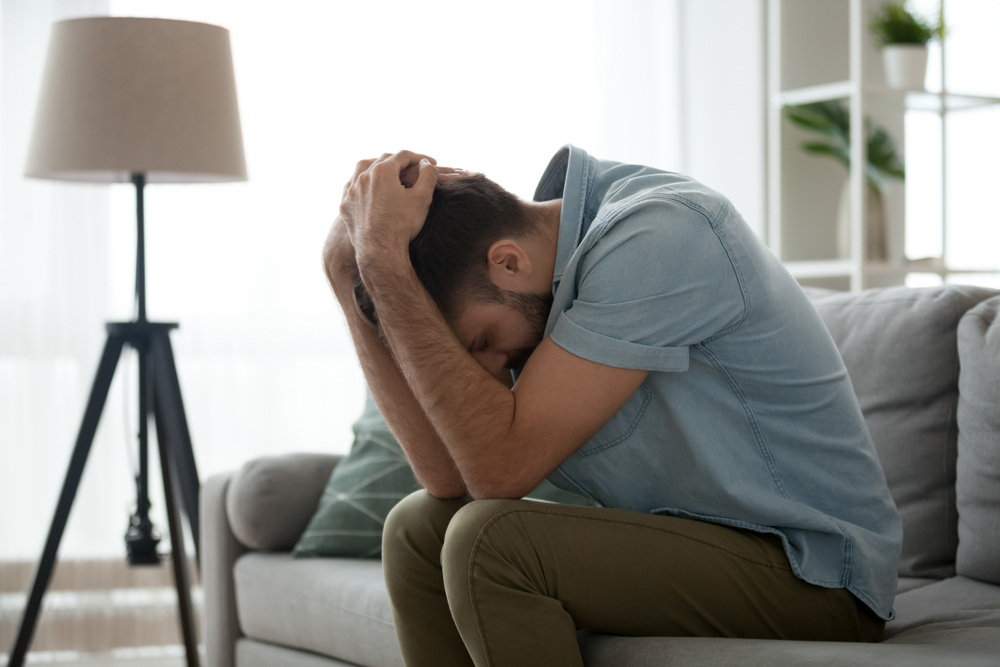 Grieving man sitting on couch holding head in hands feeling pain lonely heartbroken upset worried hopeless